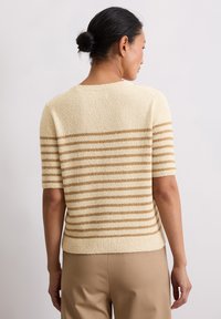 Sweter dziany w kolorze kremowym z złotymi paskami, krótkimi rękawami, ściągaczem u dołu i teksturowaną tkaniną. Modelka pokazana od tyłu, ubrana w beżowe spodnie.