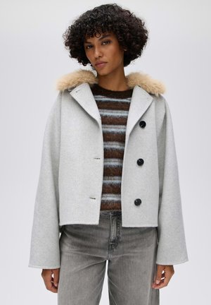 Donna che indossa un cappotto corto grigio chiaro con collo in pelliccia e bottoni neri sopra un maglione peloso a righe marroni e blu e jeans grigi.