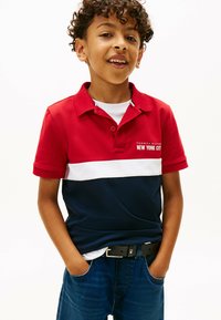 Rød, hvid og marineblå stribet polo skjorte med krave og logo, parret med mørkeblå denim jeans og et sort bælte. Korte ærmer.