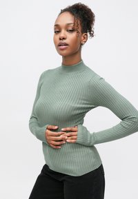 Πλεκτό ριμπ ζιβάγκο σε ανοιχτό πράσινο. Διαθέτει μακριά μανίκια με τρύπες για τον αντίχειρα, κατασκευασμένο από απαλό, υφασμάτινο ύφασμα με υφή. Καμπυλωτό τελείωμα.