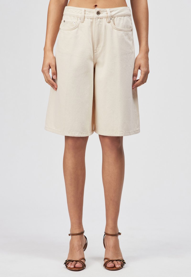 Vrouw draagt een crèmekleurige, knielange korte broek met hoge taille en voorzakken, gecombineerd met bruine sandalen met hak en bandjes, en staat tegen een effen achtergrond.