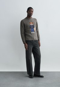 Πουλόβερ σε ανάγλυφη γκρι πλέξη με σχέδιο αλεπού σε ριγέ πουκάμισο, συνδυασμένο με φαρδιά μαύρα τζιν και μαύρα παπούτσια. Ουδέτερο φόντο.