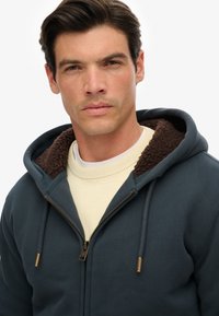 Man in een donkerblauwe rits hoodie met bruine fleecelinning en een crèmekleurige trui eronder tegen een simpele witte achtergrond.