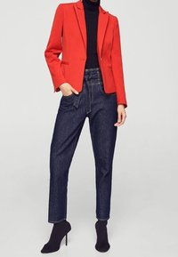 Blazer rosso dal design sartoriale, abbinato a jeans in denim scuri a vita alta. Maglione nero a collo alto sotto. Stivaletti neri alla caviglia completano il look.