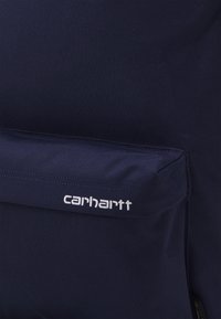Tmavě modrá látková batohová kapsa s bílým vyšitým logem Carhartt umístěným uprostřed klopy kapsy.