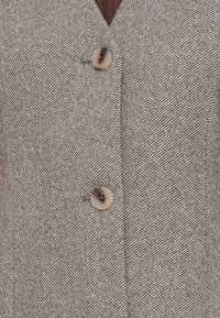 Blazer de color marrón claro con un patrón de espiga, dos botones grandes y un diseño de cuello en V.