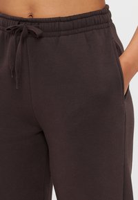 Tumši brūni sporta bikses, kas izgatavotas no mīksta materiāla, ar elastīgu jostu un auklu, sānu kabatām un brīvu piegriezumu.