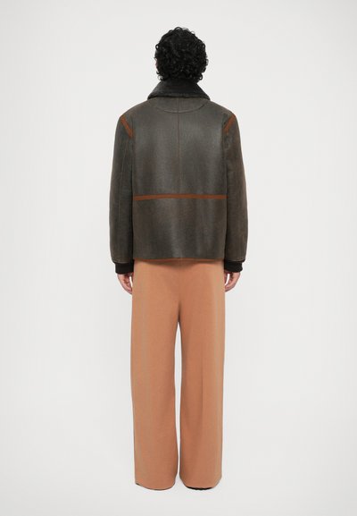 Brown shearling jack met een hoge kraag, voorzien van contrasterende oranje stiksels, en relaxte beige broeken. Zachte textuur zichtbaar.