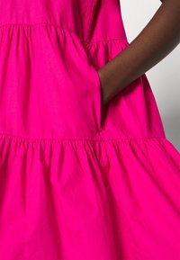 Vestido fucsia con un diseño en capas, que cuenta con un bolsillo y una textura suave. La tela parece ligera y aireada.