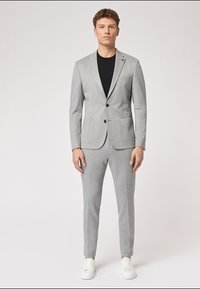 Hellgrauer Anzug mit tailliertem Blazer und schmal zulaufenden Hosen, kombiniert mit einem schwarzen T-Shirt und weißen Sneakern. Weicher Stoff, klassisches Design.