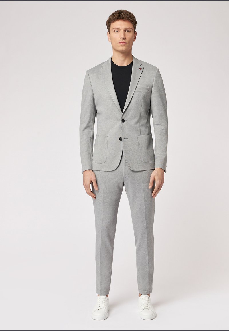 Hellgrauer Anzug mit tailliertem Blazer und schmal zulaufenden Hosen, kombiniert mit einem schwarzen T-Shirt und weißen Sneakern. Weicher Stoff, klassisches Design.