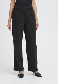 Donna che indossa pantaloni neri a gamba larga con un piccolo motivo a pois e décolleté neri con punta appuntita e fibbie su uno sfondo bianco.