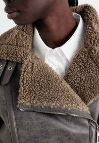 Grå läderjacka med en bred, brun fårskinnskrage. Har dragkedjestängning och en texturerad finish som kompletterar en vit skjorta med krage.
