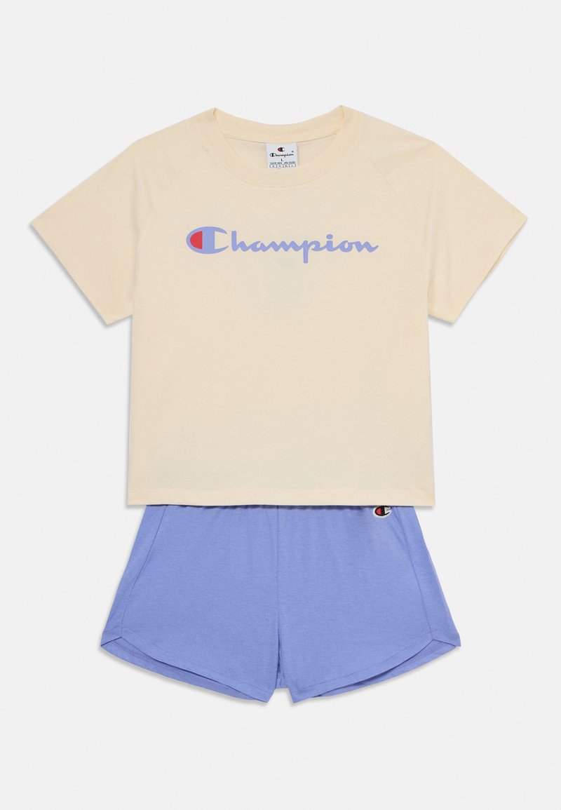 Gaiši beidžu Champion T-krekls ar purpura krāsas logo, kombinēts ar ziliem sporta šortiem, kuru jostā ir neliels logo. Mīksts audums, ikdienišķs dizains.