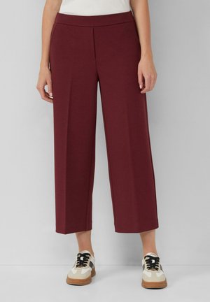 Pantalons larges bordeaux en tissu lisse, avec un devant plat et une taille élastique, associés à des baskets beiges et noires.