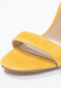 Sandale ouverte jaune en daim avec une bride avant unique et des détails cousus sur un fond clair.