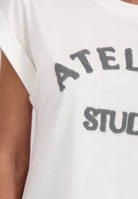 Witte blouse met korte mouwen, versierd met verhoogde grijze tekstletters die gedeeltelijk de woorden "ATEL" en "STUD" spellen.