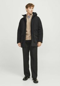 Parka negra con capucha, cierre frontal de cremallera y botones a presión, dos grandes bolsillos frontales, usada sobre un suéter beige y pantalones oscuros.