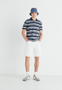 Paita polo garis-garis berwarna navy dan putih, dipadukan dengan celana pendek putih. Topi bucket dan sepatu sneakers melengkapi penampilan. Tekstur halus, potongan kasual.