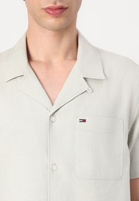 Hellgraues Kurzarmshirt aus strukturiertem Material. Es hat einen spitzen Kragen, eine Knopfleiste vorne und eine Tasche mit einem kleinen Logo in rot, weiß und blau.