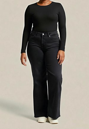 Person trägt ein schwarzes langärmeliges Oberteil, schwarze hoch taillierte weit geschnittene Jeans und weiße Schuhe, steht vor einem einfarbigen Hintergrund.