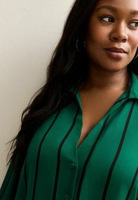 Blouse verte boutonnée avec des rayures horizontales noires. Dotée d'un col et d'un tissu lisse et léger. Manches longues et coupe décontractée.