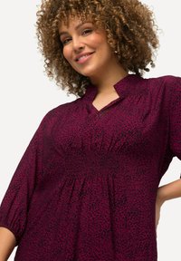 Lila Bluse mit geraffter Taille, hochgeschlossenem Kragen und dreiviertellangen Puffärmeln. Der Stoff hat ein gepunktetes schwarzes Muster.
