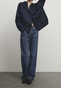 Chemise à boutons bleu marine avec manches larges associée à un jean bleu ample. L'ensemble présente un design minimaliste et des textures lisses.