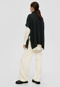 Donkergrijze gebreide poncho met een hoge kraag, voorzien van zijsplitten; gedragen over een lichtkleurig, geknopt shirt en crèmekleurige ribbroek.
