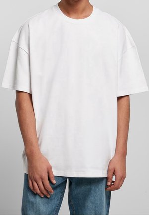 Persona con una camiseta blanca lisa de manga corta y de gran tamaño, y jeans azules, de pie frente a un fondo gris neutro.