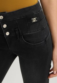 Jeans neri in denim a vita alta con chiusura a tre bottoni, accessori in argento e un piccolo dettaglio del logo dorato vicino alla vita.