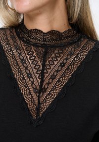 Czarna bluzka z misternym czarnym koronkowym dekoltem o falbaniastych krawędziach, prezentująca szczegółowy wzór na tle jednolitego materiału.