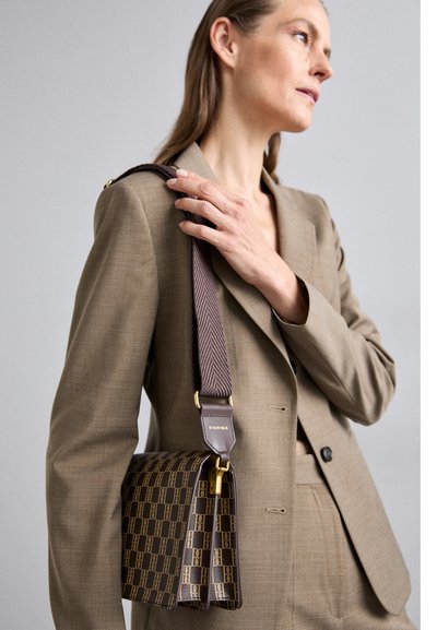 Brown patterned handbag with a structured design, featuring a herringbone strap and gold hardware accents. Worn over a beige suit jacket.