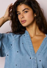 Šviesiai mėlyna džinsinė palaidinė laisvo kirpimo, su mažais juodais siuvinėjimais širdelės formos, V formos iškirpte ir subtiliu blizgesiu. Auksinė kaklo grandinėlė kaip akcentas.