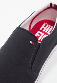 Marinblå canvas slip-on skor med vit gummisula. Har en röd och vit logotyp inuti och en liten tri-färgad flaggdetalj nära elastiksidan.