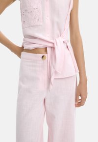 Jasnoróżowa lniana bluzka z haftowanym kwiatowym wzorem i zapięciem na guziki; zestawiona z pasującymi lekkimi spodniami z drewnianym guzikiem.