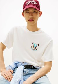 Witte katoenen t-shirt met een grafische print van "NYC" met blauwe en bruine accenten, gecombineerd met een lichtblauwe en witte gestreepte knopenhemd om de taille.