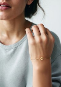 Bracelet en chaîne dorée avec un charme hexagonal, porté sur une main, sur un fond gris clair. Texture subtile et design délicat.