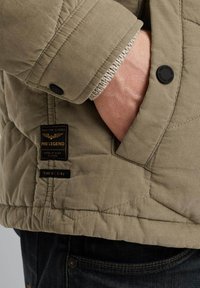 Chaqueta acolchada en verde oliva con textura de canalé, que presenta una etiqueta de parche y un botón a presión. Los puños muestran un detalle interior de punto.