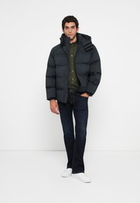 Mand iført sort pufferjakke, olivengrøn skjorte med knapper, mørke jeans og hvide sneakers, stående mod en hvid baggrund.