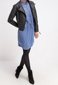 Černá kožená motorkářská bunda s prošívanými detaily na ramenou, nosená přes světle modré džíny šaty, doplněná černými kotníkovými botami a legínami.