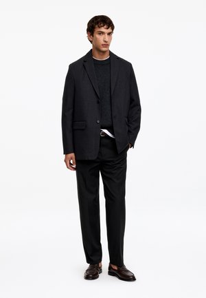 Homme debout portant un blazer noir, un pull foncé sur une chemise blanche, un pantalon noir et des mocassins marron foncé sur fond blanc.