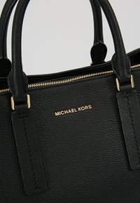 Sac à main en cuir noir texturé avec double poignée, fermeture éclair dorée et logo "MICHAEL KORS" en lettres dorées sur le devant.