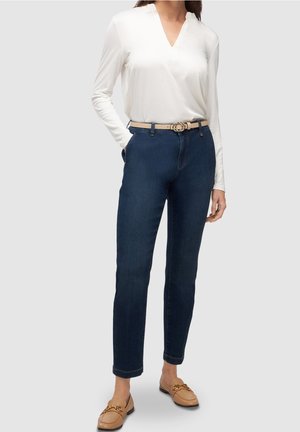 Frau trägt weiße Langarmbluse, dunkelblaue Jeans mit beigem Gürtel und beige Loafers, steht mit einer Hand in der Tasche.