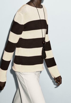 Person wearing black and white wide striped knit sweater with long black tassel necklace and white pants against a plain light background.