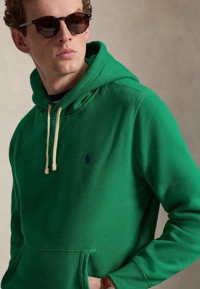 Jeune homme portant des lunettes de soleil marron et un sweat à capuche vert avec des cordons beige, debout devant un fond beige uni.