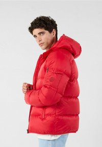 Giacca a vento rossa con cappuccio, chiusura frontale con zip e sezioni imbottite. Dotata di tasca sulla manica con dettaglio logo. Texture liscia e lucida.