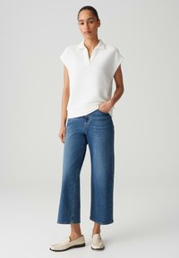 Polo bianca a maniche corte in tessuto testurizzato abbinata a jeans blu a gamba larga e mocassini crema. Outfit semplice e informale.