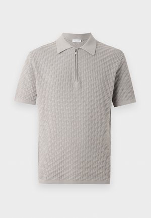 Άνδρας που φοράει ανοιχτό γκρι πικέ πόλο με κοντά μανίκια, χακί παντελόνι και μαύρα δερμάτινα μοκασίνια, όρθιος με τα χέρια στις τσέπες.