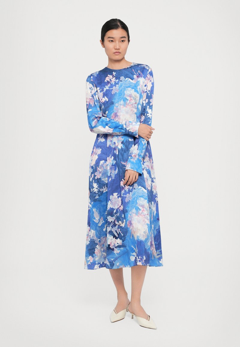 Vrouw in een lange mouwen, blauwe bloemen midi-jurk en witte schoenen met een lage hak, staand tegen een effen lichte achtergrond.